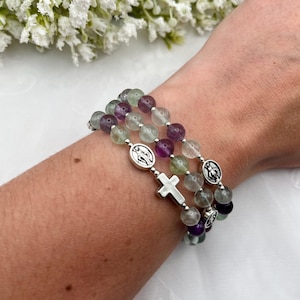 May include: A multi-strand bracelet featuring smooth, round beads in shades of translucent green and purple. The bracelet is accented with silver-toned metal charms, including two oval-shaped religious medallions and a small, plain cross charm.