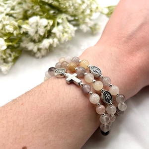 May include: Three beaded bracelets with neutral-toned beads are displayed. Each bracelet features a silver cross and religious medallions. The bracelets are worn on a wrist, with a soft background of white and green.