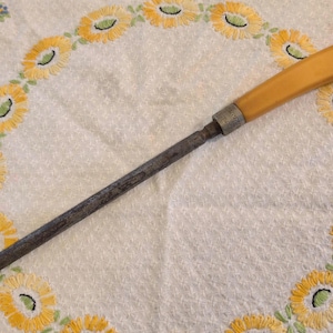 Op de afbeelding: Een antieke messenslijper met een lichtbruin handvat en een donkere metalen slijpstaaf. Het handvat is rechthoekig en de staaf loopt taps toe naar een punt. Het ligt op een witte doek met gele bloemenborduurwerk.