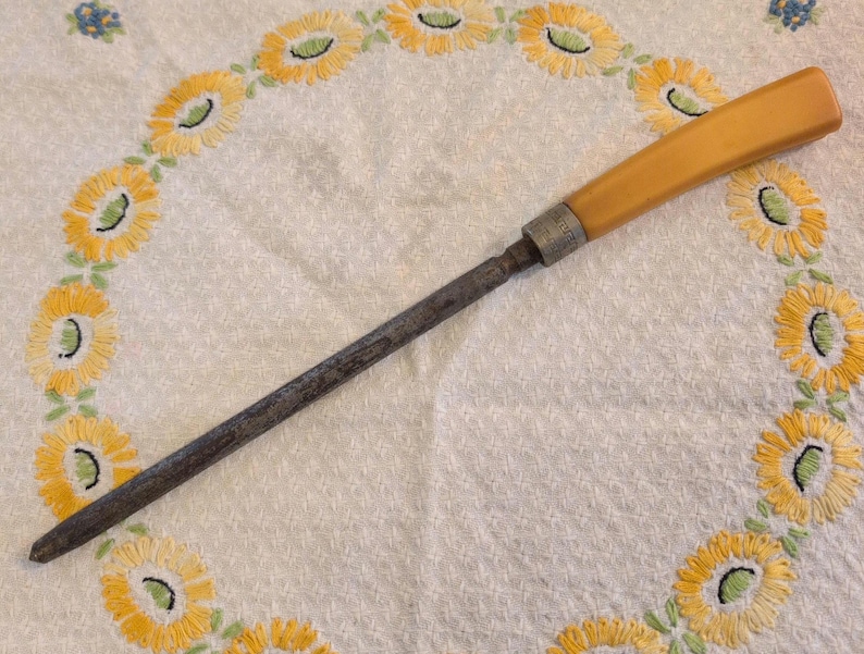 Op de afbeelding: Een antieke messenslijper met een lichtbruin handvat en een donkere metalen slijpstaaf. Het handvat is rechthoekig en de staaf loopt taps toe naar een punt. Het ligt op een witte doek met gele bloemenborduurwerk.