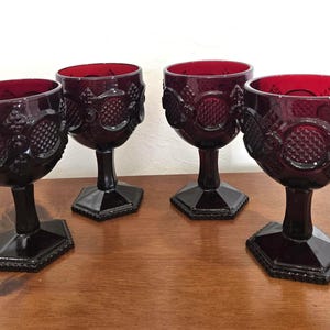 Peut inclure: Quatre gobelets rouge rubis avec des motifs ornés et texturés. Les gobelets présentent un motif détaillé autour du bol et reposent sur une base hexagonale foncée. La tige et la base sont d'une couleur foncée et unie, et les bols sont remplis de la même couleur rouge rubis.