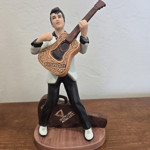 May include: A ceramic figurine of Elvis Presley playing a guitar. The figure is wearing a white jacket, black trousers, and white shoes. The guitar is brown with decorative carvings and the words "ELVIS PRESLEY". A brown case with "MEMPHIS" is at his feet.
