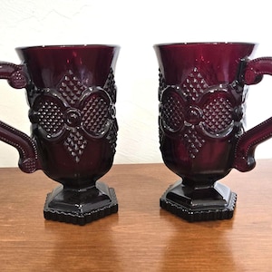 Könnte beinhalten: Zwei rubinrote Glasbecher mit verzierten Griffen und detaillierten geprägten Designs. Die Becher haben einen Fuß und ein dekoratives Muster am Körper. Die Becher sind wahrscheinlich Vintage oder antik und eignen sich zum Servieren von Getränken.