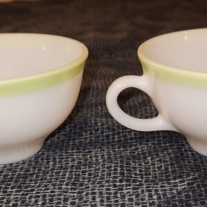 May include: Two white teacups with a pale green band around the rim. Each cup has a curved handle and a rounded base. The cups are set against a dark, textured background.