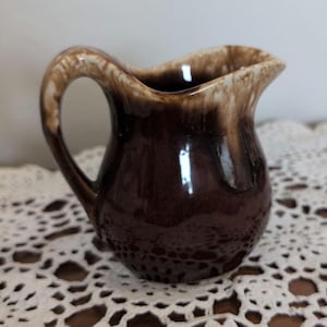 May include: A small, vintage brown ceramic creamer pitcher with a curved handle. The pitcher has a glossy finish with a two-tone color scheme, featuring a darker brown base and a lighter brown rim. The creamer is sitting on a white lace doily.