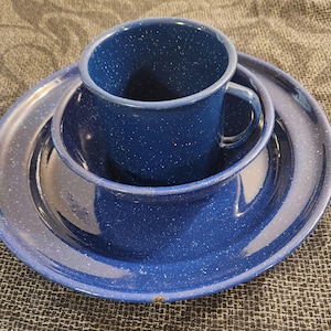 May include: A blue speckled enamelware camping dish set. The set includes a cup with a handle, a bowl, and a plate. The blue colour has white speckles throughout. The set is designed for outdoor use.