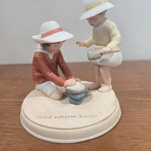 May include: A ceramic figurine depicting two children playing with a bucket. One child kneels, wearing a red sweater and white hat, while the other stands, wearing a yellow sweater and white hat. The base is inscribed with "Jessie Willcox Smith."