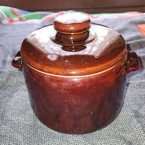 May include: A brown ceramic crock pot with a lid. The crock pot has a white rim around the top and a knob on the lid.