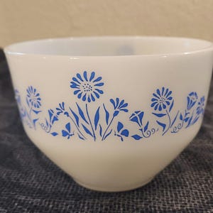 Op de afbeelding: Witte keramische mengkom met een blauw bloemenpatroon rond de rand. De bloemen zijn gestileerd en omvatten madeliefjes en andere kleine bloemen.