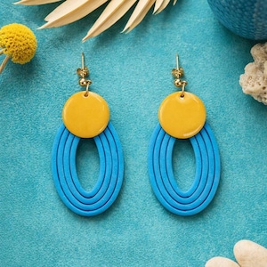 Könnte beinhalten: Ein Paar handgefertigte Ohrringe mit modernem Design. Jeder Ohrring hat einen gelben Kreis und eine blaue, geschichtete ovale Form. Die Ohrringe hängen an goldfarbenen Ohrhaken und bilden einen auffälligen Kontrast.