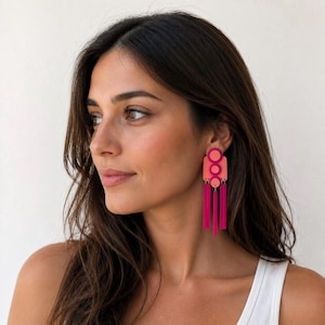May include: Bright pink and coral earrings with a geometric design. The earrings have a circular top, a rectangular middle, and three long, dangling tassels. A striking accessory, these earrings are a vibrant statement piece.