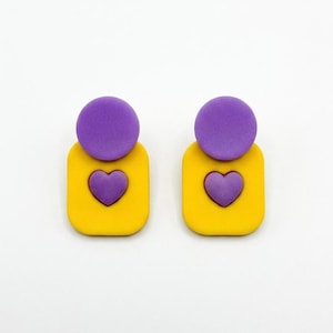 Peut inclure: Paire de boucles d'oreilles faites à la main. Chaque boucle d'oreille est composée d'un cercle violet sur un carré jaune aux coins arrondis. Un cœur violet est au centre du carré jaune, sur fond blanc.
