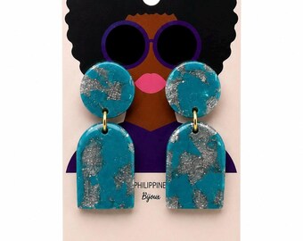 May include: Teal and silver-flecked dangle earrings featuring a circular top piece connected to a larger arch-shaped bottom piece by a gold-toned metal ring. The earrings are displayed on a card with an illustration of a person wearing sunglasses.