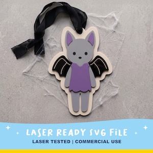 May include: A layered wooden cutout of a grey bat character with purple inner ears and a purple dress, featuring black wings and a black ribbon hanger. The text LASER READY SVG FILE and LASER TESTED | COMMERCIAL USE is printed at the bottom.