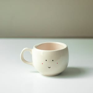 May include: A cream-coloured ceramic mug with a rounded body and a small handle. The mug has a hand-drawn face with black dots for eyes, red dots for cheeks, and a curved black line for a smile. The interior is a pale pink.