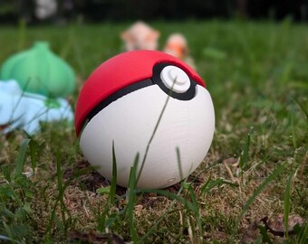 May include: A red and white Poké Ball sits in the grass, with a black band and white button detail. A blue and green Bulbasaur figure is in the background. The scene is set outdoors in a grassy area.