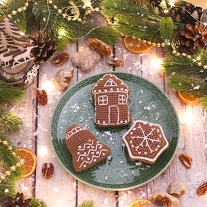 May include: A festive arrangement featuring a green plate with three crocheted Christmas ornaments: a gingerbread house, a bell, and a snowflake. The ornaments are brown with white detailing. The scene includes pine branches, lights, and dried orange slices.