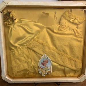 Può includere: Un abito di raso giallo e pizzo in una scatola di presentazione in plastica trasparente. L'abito è piegato a metà e ha un bordo in pizzo. La scatola ha un'etichetta con il nome "Julia" su di essa.
