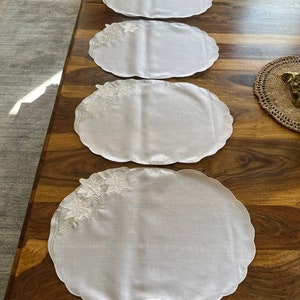 May include: Four white oval-shaped placemats with scalloped edges and embroidered floral designs in a light green color. The placemats are arranged on a wooden table.