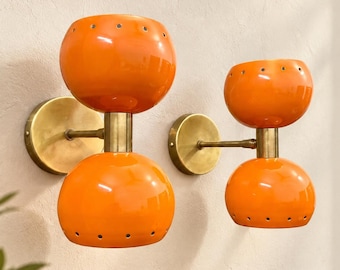 May include: Two wall sconces featuring dual orange glass globes with small circular perforations at the top and bottom edges. The globes are connected by a central brass-toned metal rod and attached to the wall with a circular brass-toned plate.
