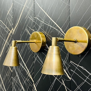 Op de afbeelding: Twee gouden wandlampen met kegelvormige lampenkappen, gemonteerd op een zwarte muur met witte lijnen.