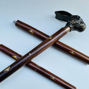 Puede incluir: Bastón decorativo con un mango en forma de cabeza de conejo. El bastón es de madera marrón oscuro con detalles dorados y una punta de latón. La cabeza de conejo es de color bronce.