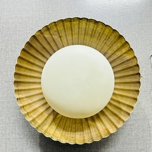 May include: A round, off-white light bulb is centered in a gold-colored, scalloped-edge bowl. The bowl has a ridged design radiating from the center. The light fixture is likely for a wall or ceiling.