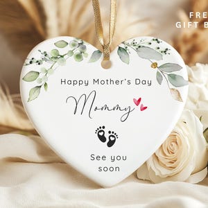 May include: A heart-shaped ceramic ornament with a gold ribbon for hanging. The ornament features the text "Happy Mother's Day Mommy" with baby footprints and floral accents. The background includes white roses and dried plants.