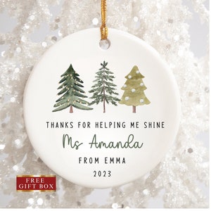K&ouml;nnte beinhalten: Wei&szlig;er Keramik-Weihnachtsbaumschmuck mit drei gr&uuml;nen Aquarellb&auml;umen, Lichtern und wei&szlig;en Schneeflocken. Der Anh&auml;nger tr&auml;gt die Aufschrift "Danke, dass du mir geholfen hast zu gl&auml;nzen Frau Amanda von Emma 2023".
