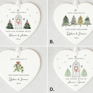 K&ouml;nnte beinhalten: Vier wei&szlig;e herzf&ouml;rmige Keramik-Ornamente mit dem Text "First Christmas at" und einem Haus mit einem Kranz an der T&uuml;r. Jedes Ornament hat eine andere Adresse und Namen. Die Ornamente sind mit Schneeflocken und Weihnachtsb&auml;umen verziert.
