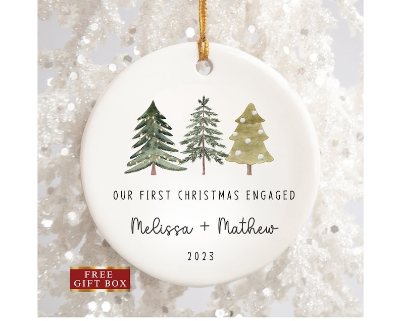 Peut inclure: Ornement de No&euml;l en c&eacute;ramique blanche avec trois arbres verts en aquarelle, deux avec des lumi&egrave;res et un avec des boules de neige blanches. Le texte indique "Our First Christmas Engaged Melissa + Mathew 2023".