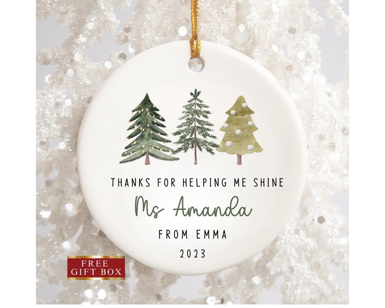 K&ouml;nnte beinhalten: Wei&szlig;er Keramik-Weihnachtsbaumschmuck mit drei gr&uuml;nen Aquarellb&auml;umen, Lichtern und wei&szlig;en Schneeflocken. Der Anh&auml;nger tr&auml;gt die Aufschrift "Danke, dass du mir geholfen hast zu gl&auml;nzen Frau Amanda von Emma 2023".