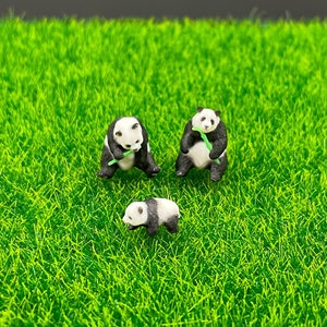 Puede incluir: Tres figuritas de panda en miniatura, dos están sentadas y sujetan un palo de bambú verde, una está de pie. Las figuritas están sobre una superficie de hierba verde.