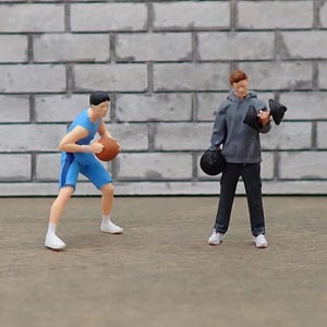 Puede incluir: Figuras en miniatura de dos personas practicando deportes. Una figura, en azul, bota un balón de baloncesto. La otra figura, con una sudadera con capucha gris, sostiene un par de guantes de boxeo negros y un saco de boxeo negro. El fondo es una pared de ladrillos gris.
