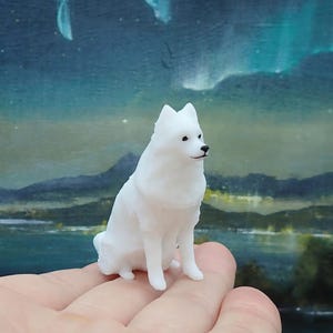 May include: A small, white figurine of a dog, possibly a Samoyed, is perched on a hand. The dog has black eyes and a black nose. The background features a landscape painting with a night sky and mountains.