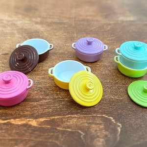 Könnte beinhalten: Ein Set aus sechs bunten Mini-Töpfen mit Deckeln. Die Töpfe sind in den Farben Rosa, Lila, Blau, Grün, Gelb und Braun. Die Deckel haben ein geriffeltes Design.
