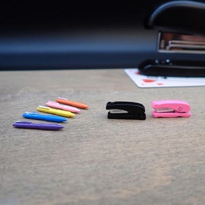 May include: A collection of colorful pens and staplers on a wooden surface. The pens are in various colors, including purple, blue, yellow, pink, and orange. There are two staplers, one black and one pink. A larger black stapler is in the background.