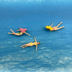 Puede incluir: Tres figuras en miniatura de personas nadando en una piscina azul. Las figuras llevan trajes de baño y tienen diferentes colores de pelo.