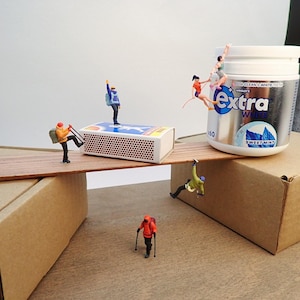 May include: A whimsical diorama featuring miniature figures interacting with everyday objects. A matchbox serves as a bridge, with figures climbing a container of "Extra" gum. The scene includes cardboard boxes and a wooden plank, creating a playful, imaginative composition.