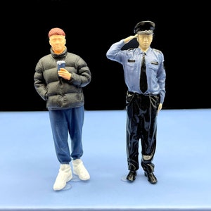 May include: Two miniature figurines, one wearing a black jacket and blue jeans, holding a cell phone, and the other wearing a blue uniform and a black hat, saluting.