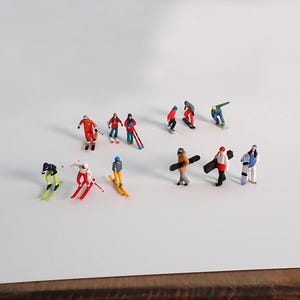 May include: Miniature skiers and snowboarders in various colorful outfits are scattered across a white surface. The figures are in action poses, some skiing, others snowboarding, and some carrying their boards. The scene evokes a winter sports theme.