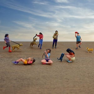 May include: A collection of miniature figurines depicting people with dogs and cats on a sandy surface, set against a cloudy sky. Some figures are walking dogs, others are playing with pets or holding them. A frisbee is being thrown.