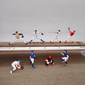 May include: Miniature baseball figurines in various action poses, including pitching, batting, and fielding. The figurines are dressed in team uniforms with different colors. They are arranged on a wooden base with a ruler marked in inches.