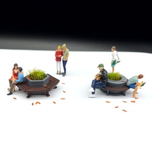 Può includere: Una scena in miniatura con sei figure sedute su due panchine esagonali con fioriere. Le panchine sono in legno e hanno una finitura marrone scuro. Le fioriere sono in plastica grigia e hanno erba verde che cresce al loro interno. Le figure indossano tutte abiti casual e sono in varie pose. La scena è ambientata su uno sfondo bianco.