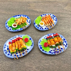 Puede incluir: Cuatro platos en miniatura con diseños azules y blancos, cada uno con un tipo diferente de comida a la brocheta. Los platos están dispuestos sobre una superficie de madera. La comida incluye camarones, pollo y verduras.