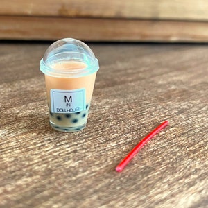 May include: A clear plastic cup with a lid filled with a peach-coloured drink and black boba pearls. The cup has a white label that reads "M ini DOLLHOUSE". A red plastic straw is lying on a wooden surface.