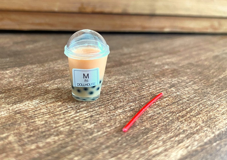 May include: A clear plastic cup with a lid filled with a peach-coloured drink and black boba pearls. The cup has a white label that reads "M ini DOLLHOUSE". A red plastic straw is lying on a wooden surface.