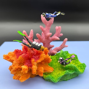 May include: Three miniature scuba divers are positioned on coral reef decorations. One diver is on a pink coral reef, while the other two are on orange and green coral reefs. The divers are wearing scuba gear and have flippers.