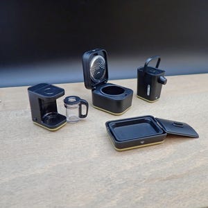 Puede incluir: Juego de electrodomésticos de cocina negros en miniatura, que incluye una cafetera, una arrocera, un hervidor y un recipiente rectangular con tapa. Los electrodomésticos tienen un ribete dorado y están sobre una superficie marrón claro.