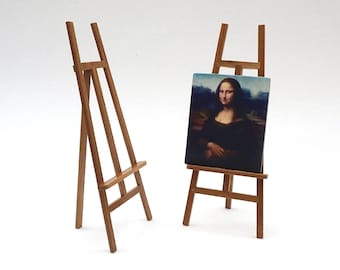 May include: Two miniature wooden easels, one empty and one holding a small painting of the Mona Lisa. The easels are made of light brown wood and stand on a white surface. The painting features the iconic portrait.